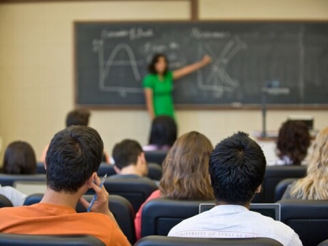 A classroom lecture in progress, with a person at the front pointing to graphs and equations on a chalkboard while seated students listen.