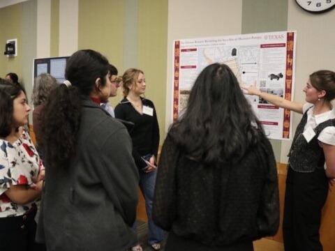 A small group gathered indoors around a research poster, with one person pointing to maps and diagrams on the display; the word ‘Texas’ is visible at the top of the poster.